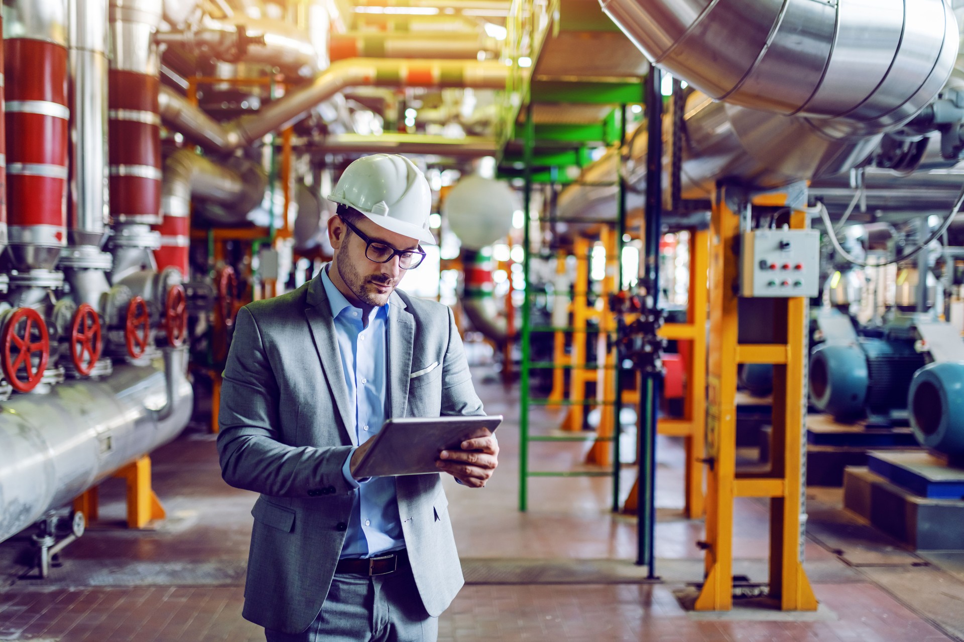 Handsome caucasian manager in gray suit and with helmet on head using tablet while standing in power plant.